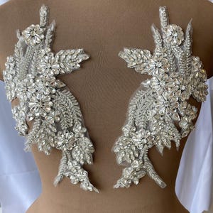 Può includere: Una coppia di applicazioni floreali simmetriche color argento con cristalli sfaccettati trasparenti, perline e paillettes disposte in motivi a foglia e fiore su una base in rete trasparente.