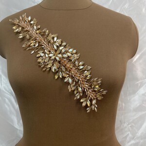 May include: A gold-toned, decorative applique featuring a linear design of clear and amber-colored crystals. The crystals are arranged in a leaf-like pattern, attached to a thin, flexible wire base. The applique is displayed on a brown mannequin.