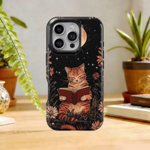 May include: A phone case featuring a tabby cat reading a book, set against a night sky with a moon and stars. The design includes flowers and foliage in a whimsical illustration style. The case is black with a glossy finish.