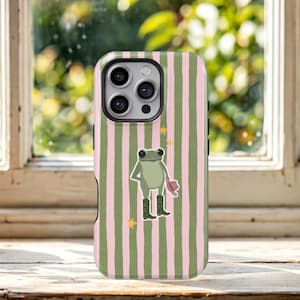 May include: A smartphone case featuring a vertical striped pattern in alternating shades of light pink and olive green. Centered on the case is an illustration of a light green frog wearing dark green boots and holding a pink hat, surrounded by small yellow stars.