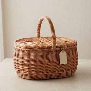 May include: An oval-shaped woven wicker basket featuring a light brown natural finish. The basket includes a hinged lid that opens in the center and a single arched handle. A small, blank, rectangular beige paper tag hangs from the handle.