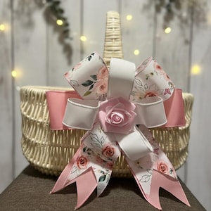 Peut inclure: Un panier en osier orné d'un nœud décoratif. Le nœud présente une rose rose au centre, un ruban blanc et un ruban rose avec un motif floral. La poignée du panier est visible.