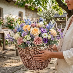Puede incluir: Una gran cesta de mimbre tejida llena de un exuberante arreglo floral con rosas de jardín en tonos melocotón, rosa y blanco, lavanda morada y flores de espuela de caballero azules. El ramo incluye pequeñas flores blancas y follaje verde.