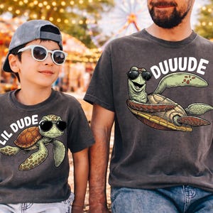 May include: Two matching charcoal gray t-shirts. One says "LIL DUDE" and the other says "DUUUDE" in white text. Each shirt features a cartoon turtle wearing sunglasses. The shirts are worn by a child and an adult.