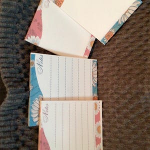 May include: Three square paper notepads featuring a white center with blue dashed lines and the word Notes printed in script. The borders are decorated with a floral pattern of white daisies with orange centers against blue and pink wavy backgrounds.