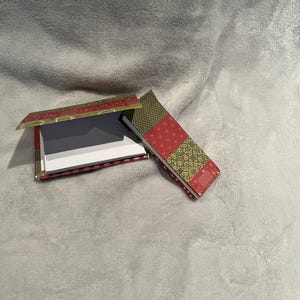 May include: Two rectangular note pads with fabric covers featuring a patchwork design of red and olive green squares. The patterns include small hearts, swirls, and dots. One pad is open to reveal white paper, while the other is closed.