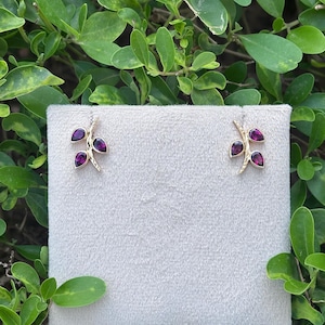 May include: A pair of gold stud earrings shaped like small branches, each featuring three teardrop-shaped deep magenta gemstones arranged along the stem. The earrings are displayed on a square, light beige, textured fabric display card.