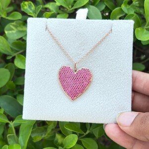 Könnte beinhalten: Eine Halskette aus Roségold mit einem herzförmigen Anhänger, der mit zahlreichen kleinen, leuchtend rosa Steinen besetzt ist. Der Anhänger hängt an einer zarten Roségoldkette und ist auf einer hellgrauen, quadratischen Schmuckkarte aus Samt befestigt.