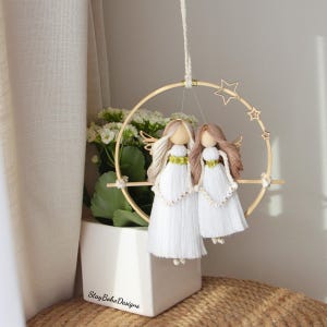 Peut inclure: Décoration suspendue faite à la main avec deux poupées anges aux longs cheveux et robes blanches. Les poupées sont suspendues dans un cercle en bois orné d'étoiles. Les poupées portent les noms "Tina" et "Asia" sur leurs robes.