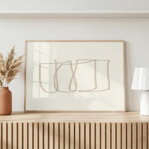 Peut inclure: Une impression d'art murale rectangulaire dans un cadre en bois clair présentant un dessin au trait beige de trois formes carrées superposées sur un fond crème. L'œuvre est exposée sur une surface en bois à lattes près d'un vase d'herbes séchées.