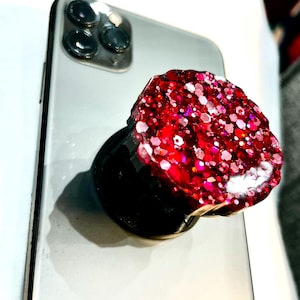 May include: A red glitter phone grip attached to a silver smartphone. The phone grip is round and covered in red and pink glitter. The phone has a black camera module.