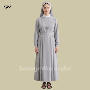 Puede incluir: Un hábito gris con cuello blanco y toca a juego. Un colgante de cruz plateada cuelga de un collar. El vestido tiene un corpiño plisado y una cintura con cinturón. El texto "Savage Wardrobe" está impreso en la parte inferior.