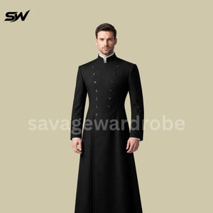 Puede incluir: Un abrigo negro de doble botonadura con cuello mandarín. El abrigo tiene varios botones y un diseño largo y fluido. El texto "savage wardrobe" está impreso en el abrigo. El abrigo lo lleva una persona.