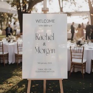 Puede incluir: Un letrero de bienvenida de boda blanco sobre un caballete de madera. El letrero dice "WELCOME to the wedding of Rachel & Morgan 15th January, 2025 THANK YOU FOR CELEBRATING WITH US!" en texto negro. El fondo muestra una recepción de boda.