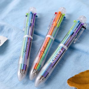 Puede incluir: Tres bolígrafos multicolores con barriles transparentes y recambios de tinta de colores arcoíris. Cada bolígrafo tiene un nombre diferente: Shawna, Justine y Kelsey. Los bolígrafos están sobre una superficie azul claro.