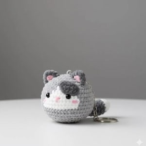 May include: A crocheted cat keychain in shades of gray, white, and black. The cat has pink accents on its ears and cheeks, with a small tail. The keychain is round and has a silver-colored metal ring.