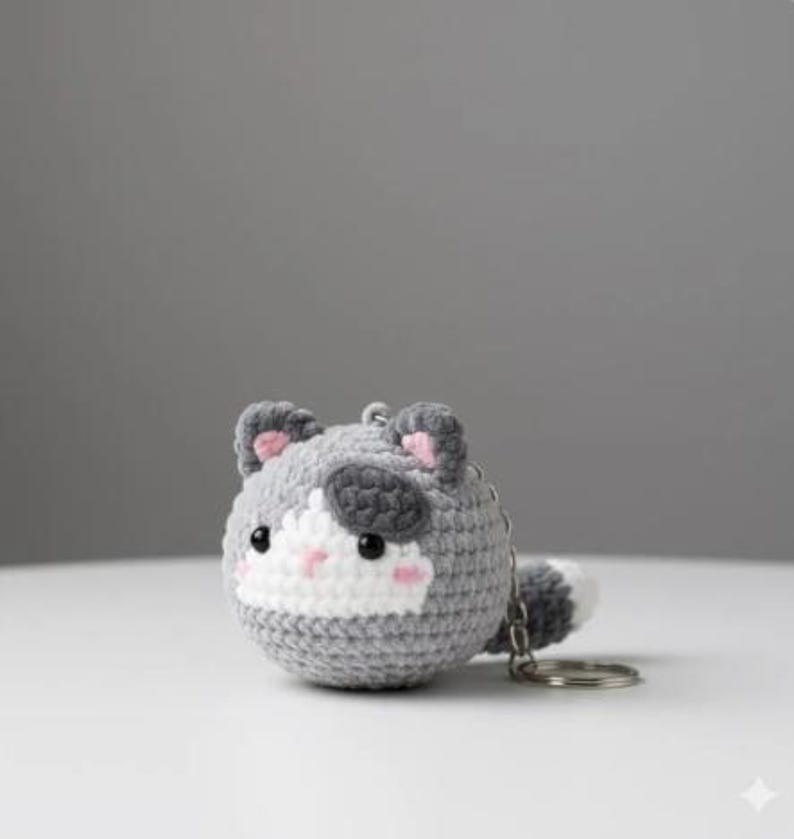 May include: A crocheted cat keychain in shades of gray, white, and black. The cat has pink accents on its ears and cheeks, with a small tail. The keychain is round and has a silver-colored metal ring.