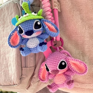 May include: Two crocheted Stitch character keychains. One is blue with a green hat, the other is pink. Both have large black eyes and are attached to pink and blue lanyards. The background is a light pink.