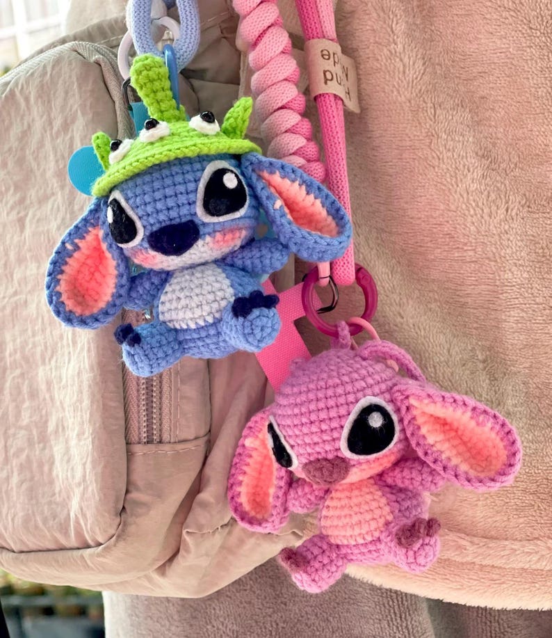 May include: Two crocheted Stitch character keychains. One is blue with a green hat, the other is pink. Both have large black eyes and are attached to pink and blue lanyards. The background is a light pink.
