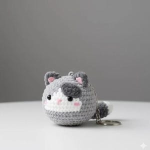 May include: A crocheted cat keyring in grey, white, and black. The cat features pink accents on its ears and cheeks, and a small tail. The keyring is round and has a silver-coloured metal ring.