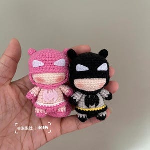 May include: Two small, handmade crochet superhero dolls. One is pink with white accents, the other is black and grey with a yellow belt. The dolls have cartoonish features and are held in a hand.