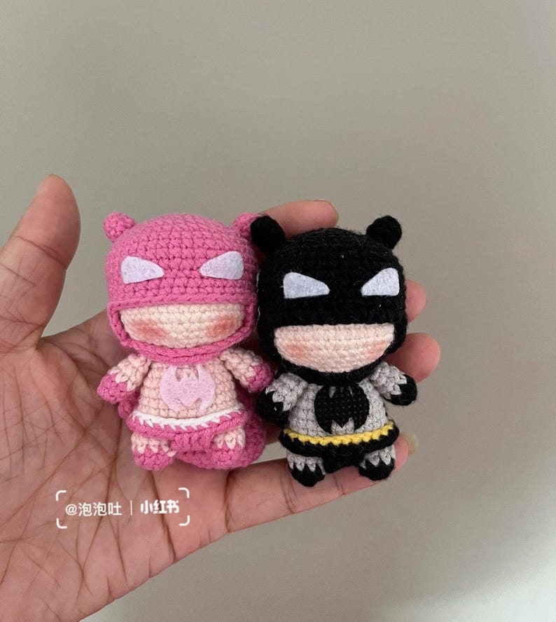 May include: Two small, handmade crochet superhero dolls. One is pink with white accents, the other is black and grey with a yellow belt. The dolls have cartoonish features and are held in a hand.