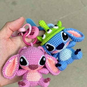 May include: Two crocheted character keychains. One is pink with large ears and a light pink inner ear. The other is blue with a green hat. Both have large black eyes and are attached to pink key rings.