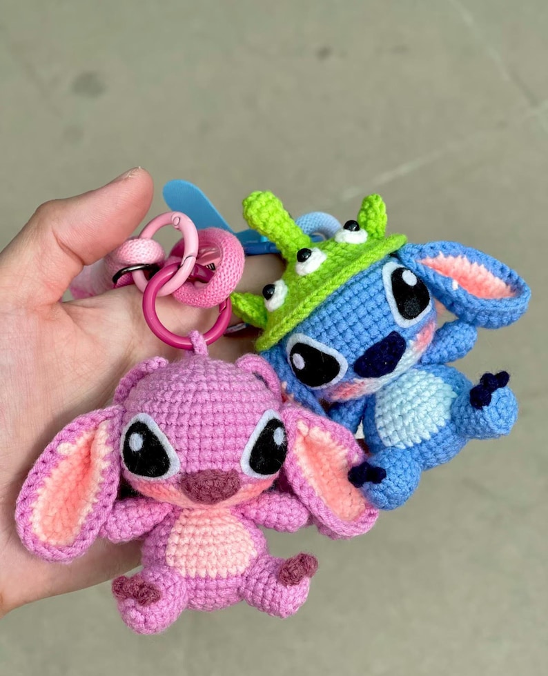 May include: Two crocheted character keychains. One is pink with large ears and a light pink inner ear. The other is blue with a green hat. Both have large black eyes and are attached to pink key rings.