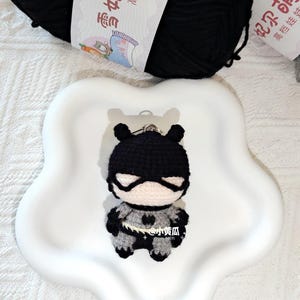 May include: A crocheted superhero keyring in black, grey, and white. The small figure has a mask and cape. The keyring is on a white, flower-shaped dish, with a black ball of yarn in the background. The figure has a heart-shaped emblem.