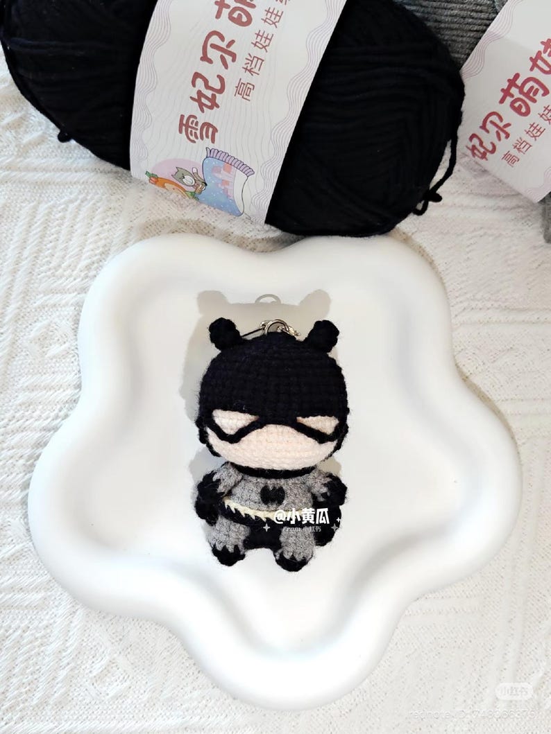 May include: A crocheted superhero keyring in black, grey, and white. The small figure has a mask and cape. The keyring is on a white, flower-shaped dish, with a black ball of yarn in the background. The figure has a heart-shaped emblem.