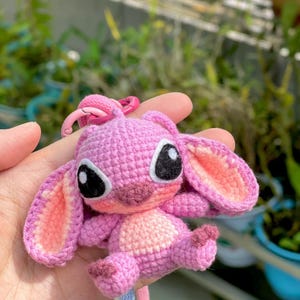 May include: A handmade pink crochet keychain featuring a cartoon character with large black eyes and pink inner ears. The keychain has a pink clasp and a blue and white striped lanyard.