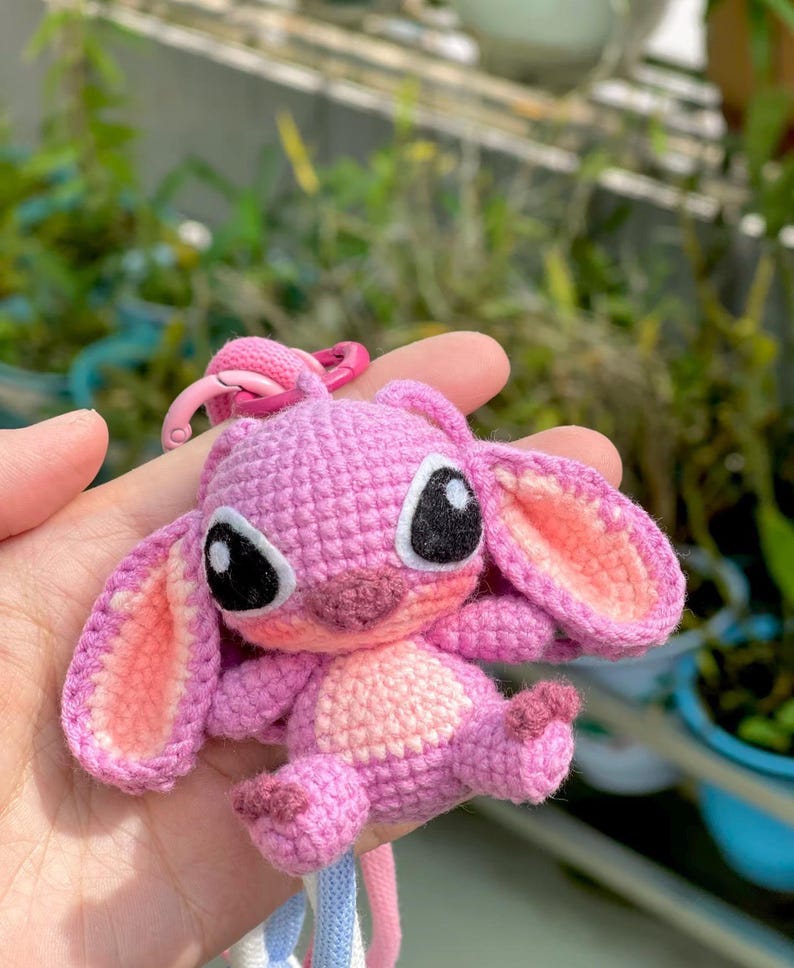 May include: A handmade pink crochet keychain featuring a cartoon character with large black eyes and pink inner ears. The keychain has a pink clasp and a blue and white striped lanyard.