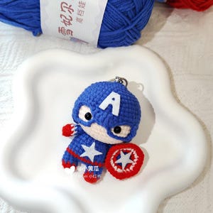 May include: A crocheted Captain America keyring. The blue and red figure features a white star and shield. The keyring sits on a white, star-shaped plate, with blue yarn visible in the background.