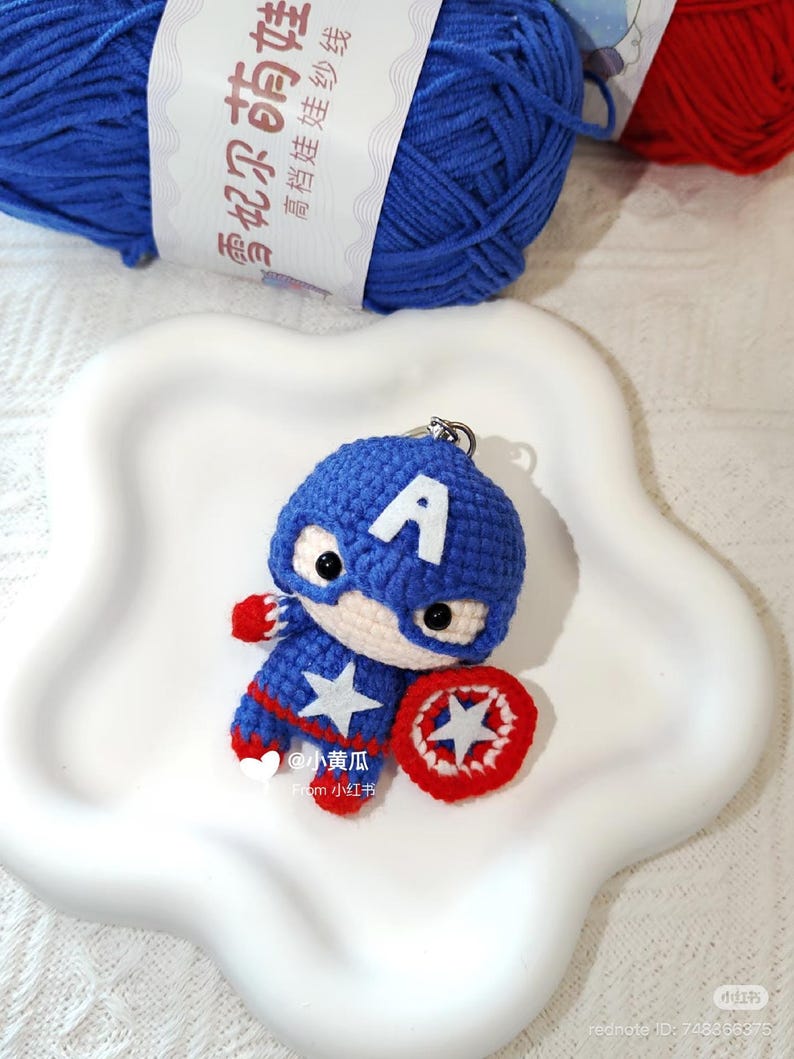 May include: A crocheted Captain America keyring. The blue and red figure features a white star and shield. The keyring sits on a white, star-shaped plate, with blue yarn visible in the background.