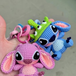 May include: Crocheted keychains featuring cartoon characters in blue, pink, and green. The blue character has large black eyes and a green hat. The pink character has large ears and black eyes. The keychains are attached to pink and blue rings.