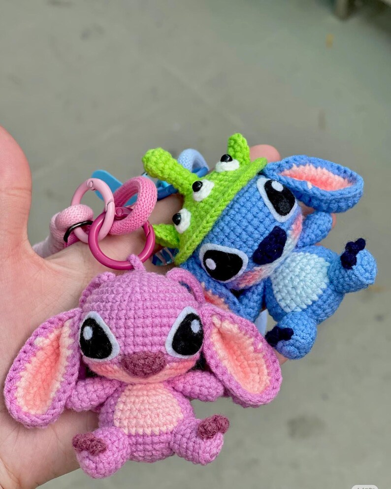 May include: Crocheted keychains featuring cartoon characters in blue, pink, and green. The blue character has large black eyes and a green hat. The pink character has large ears and black eyes. The keychains are attached to pink and blue rings.
