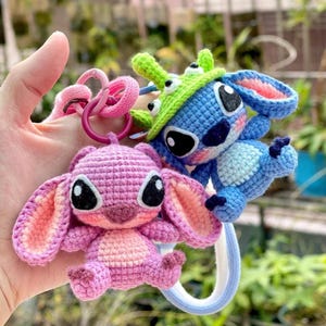 May include: Crocheted keychains featuring a blue and a pink character with large ears and black eyes. The blue character wears a green hat. The keychains are attached to pink and purple rings.