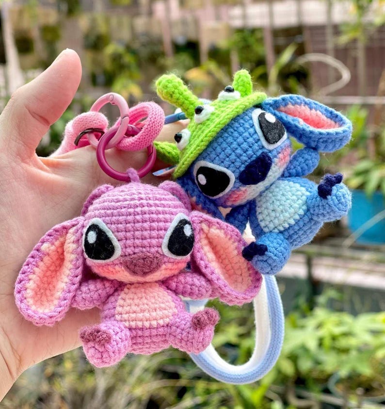 May include: Crocheted keychains featuring a blue and a pink character with large ears and black eyes. The blue character wears a green hat. The keychains are attached to pink and purple rings.