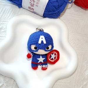 May include: A crocheted Captain America keyring in blue, red, and white yarn. The figure has a shield, a star, and the letter "A" on the head. The keyring is displayed on a white, star-shaped plate, with blue yarn visible in the background.