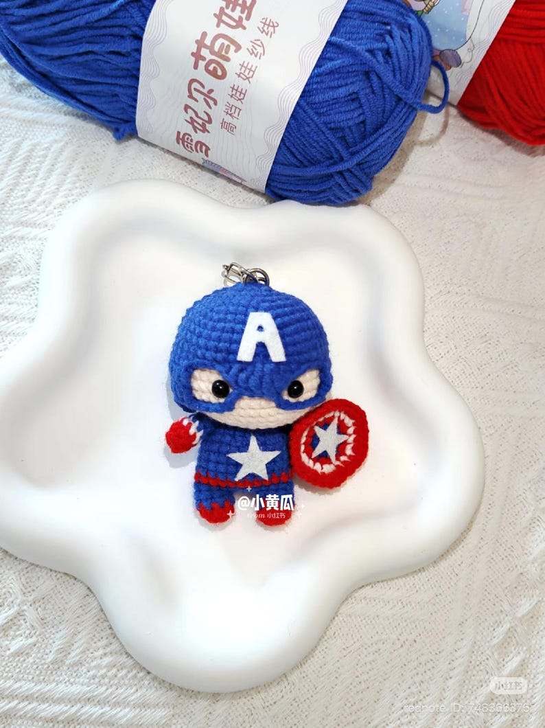 May include: A crocheted Captain America keyring in blue, red, and white yarn. The figure has a shield, a star, and the letter "A" on the head. The keyring is displayed on a white, star-shaped plate, with blue yarn visible in the background.