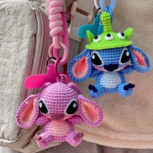 May include: Two handmade crochet keychains. One is a pink character with large ears, the other is blue with a green hat. Both have black eyes and are attached to pink and blue lanyards.