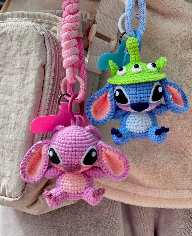 May include: Two handmade crochet keychains. One is a pink character with large ears, the other is blue with a green hat. Both have black eyes and are attached to pink and blue lanyards.