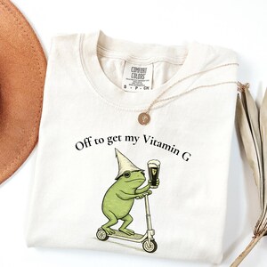 May include: Cream-colored t-shirt with the text "Off to get my Vitamin G." A cartoon frog wearing a hat and sunglasses is riding a scooter and holding a pint of beer. A gold necklace and a brown hat are also in the image.