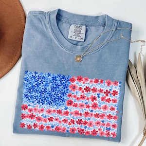 Puede incluir: Una camiseta azul con un dise&ntilde;o floral de la bandera estadounidense. La bandera est&aacute; hecha de flores azules y rojas. Un collar dorado con un colgante est&aacute; sobre la camiseta. La camiseta est&aacute; doblada sobre una superficie blanca.