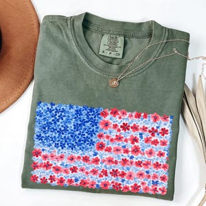 Puede incluir: Camiseta verde oliva con un dise&ntilde;o floral de la bandera estadounidense en rojo, blanco y azul. La camiseta tiene cuello redondo y mangas cortas. Un collar dorado con un colgante est&aacute; drapeado sobre la camiseta.