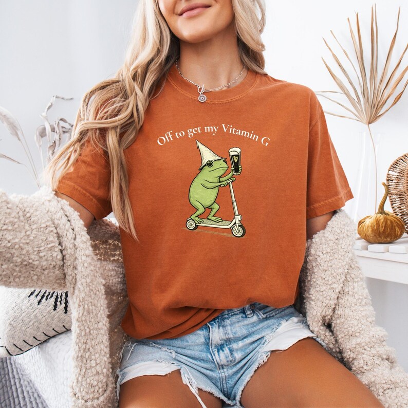 May include: Burnt orange t-shirt with the text "Off to get my Vitamin G." The graphic shows a green frog wearing a hat and sunglasses, riding a scooter while holding a pint of beer. The shirt is a comfortable, casual style.