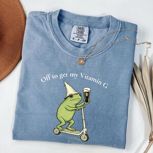 May include: A light blue Comfort Colors t-shirt with the phrase "Off to get my Vitamin G" and a graphic of a green frog riding a scooter and holding a beer. A gold necklace and a brown hat are also visible.