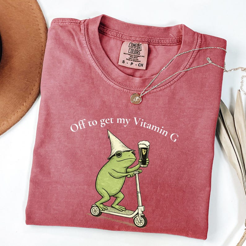 May include: A dusty rose Comfort Colors t-shirt with the phrase "Off to get my Vitamin G" and a cartoon frog riding a scooter while holding a pint of beer. A silver necklace with a small pendant is draped across the shirt.