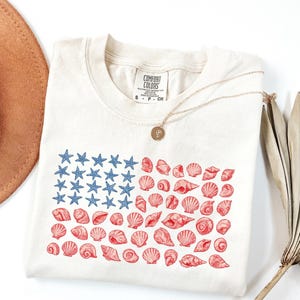May include: Cream-colored t-shirt featuring a unique American flag design. The flag's stars are blue, and the stripes are composed of red seashell illustrations. A gold necklace with a pendant rests on the shirt.