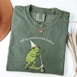 May include: An olive green t-shirt with the phrase "Off to commit Irish foolery" and a cartoon frog riding a scooter. The frog wears a white hat and holds a four-leaf clover. A gold necklace and a brown hat are also visible.